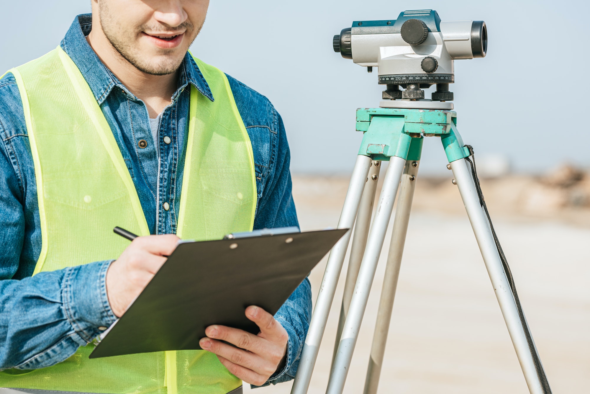 Cropped shot of smiling surveyor writing on clipboard beside digital level